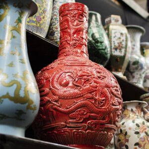 Vaso vermelho com relevo de dragão em destaque entre outros vasos ornamentais em uma prateleira.