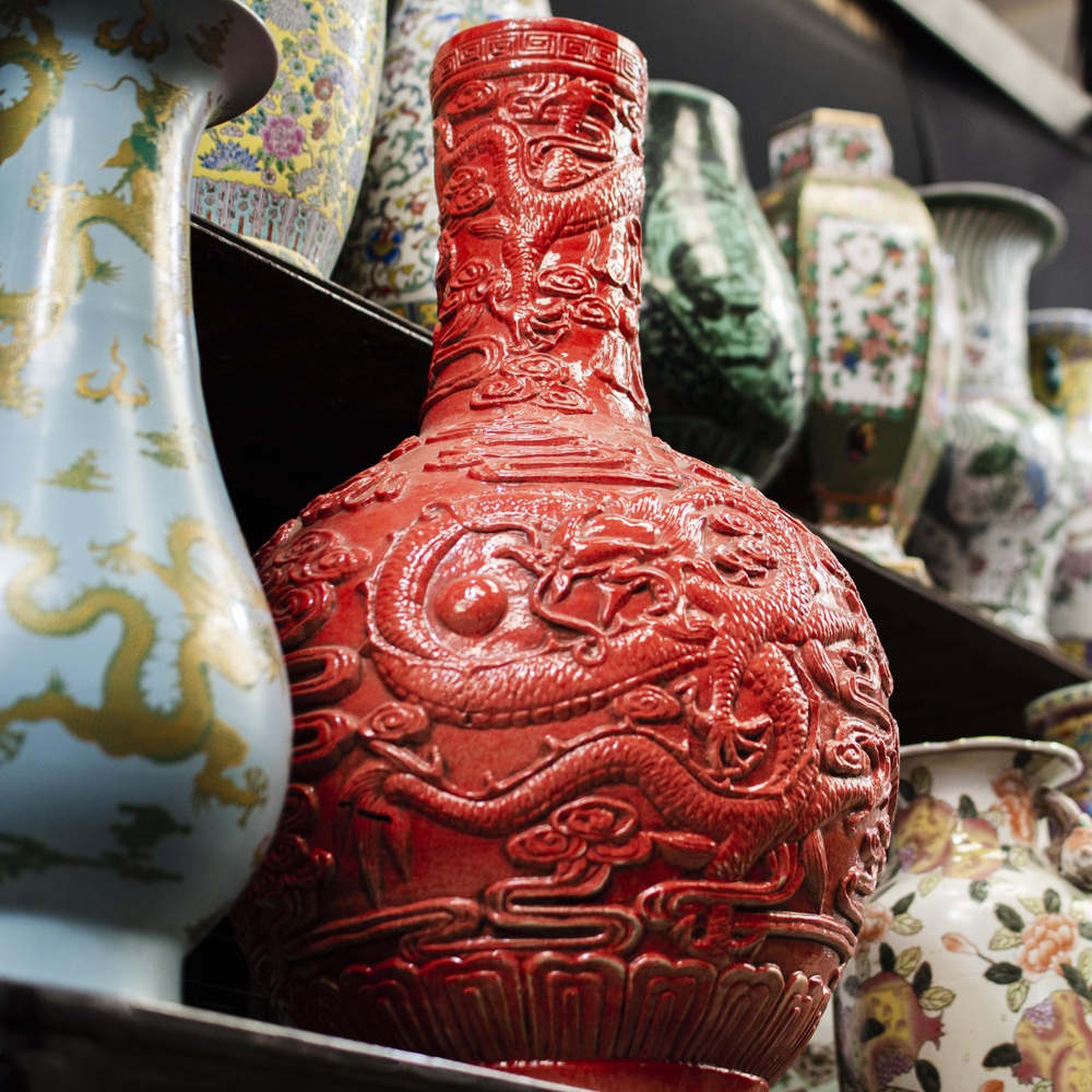 Vaso vermelho com relevo de dragão em destaque entre outros vasos ornamentais em uma prateleira.